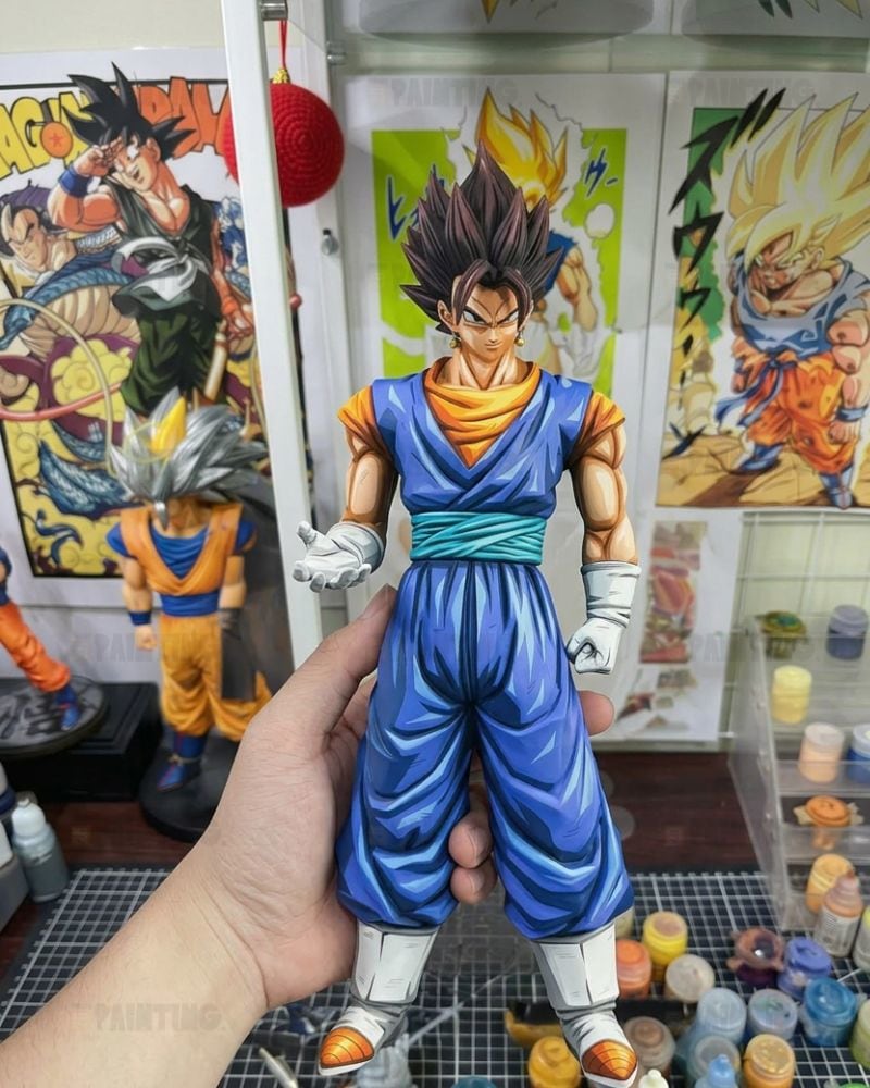 A hand holds a detailed anime action figure of Gohan from Dragon Ball Z, wearing his blue gi with an orange undershirt and white gloves, set against a background of Dragon Ball posters and art supplies.