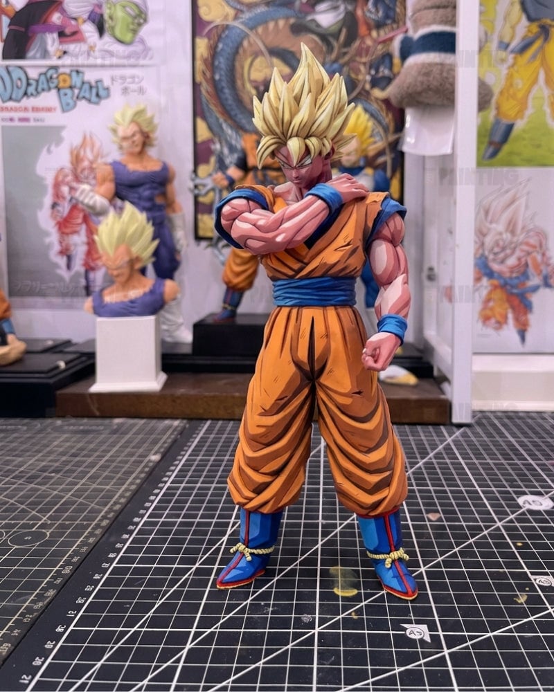 A detailed anime figurine of Goku in a Super Saiyan pose, standing on a black gridded cutting mat with Dragon Ball themed posters and other collectibles in the background.