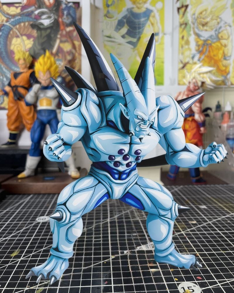 A detailed light blue and white Dragon Ball Z anime figure of a spiky monster, posed on a cutting mat with other collectible figures in a hobby room background.