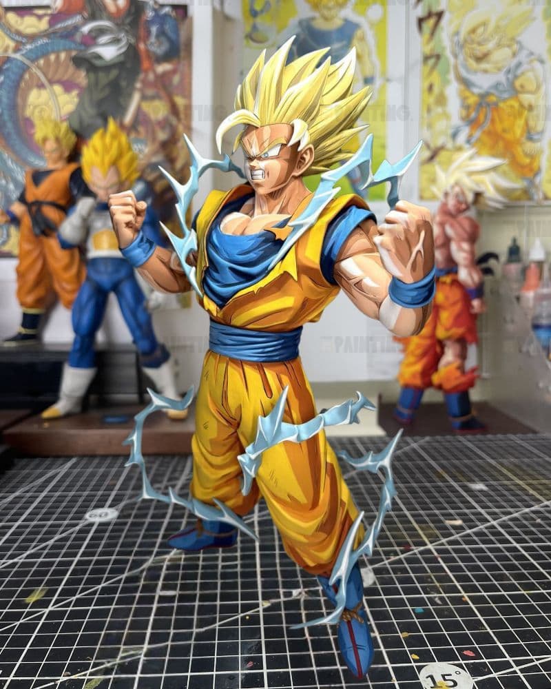 A detailed anime figurine of Goku in a Super Saiyan form, standing on a gridded cutting mat inside a hobby room with other Dragon Ball collectibles in the background.
