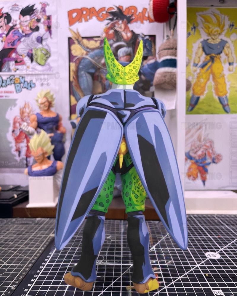 A Dragon Ball figure of Cell from the back, featuring large blue and black wing-like back plates, green textured armor, and yellow boots, displayed on a cutting mat with Dragon Ball posters in the background.