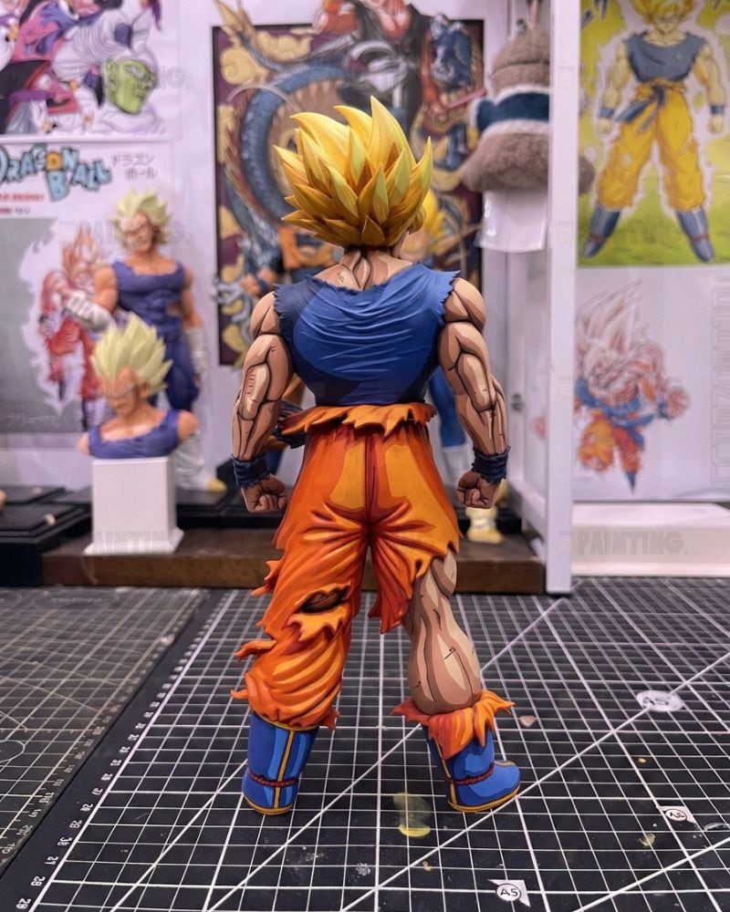 A detailed anime action figure of Goku in a Super Saiyan form, shown from the back on a black gridded cutting mat with other collectibles in the background.