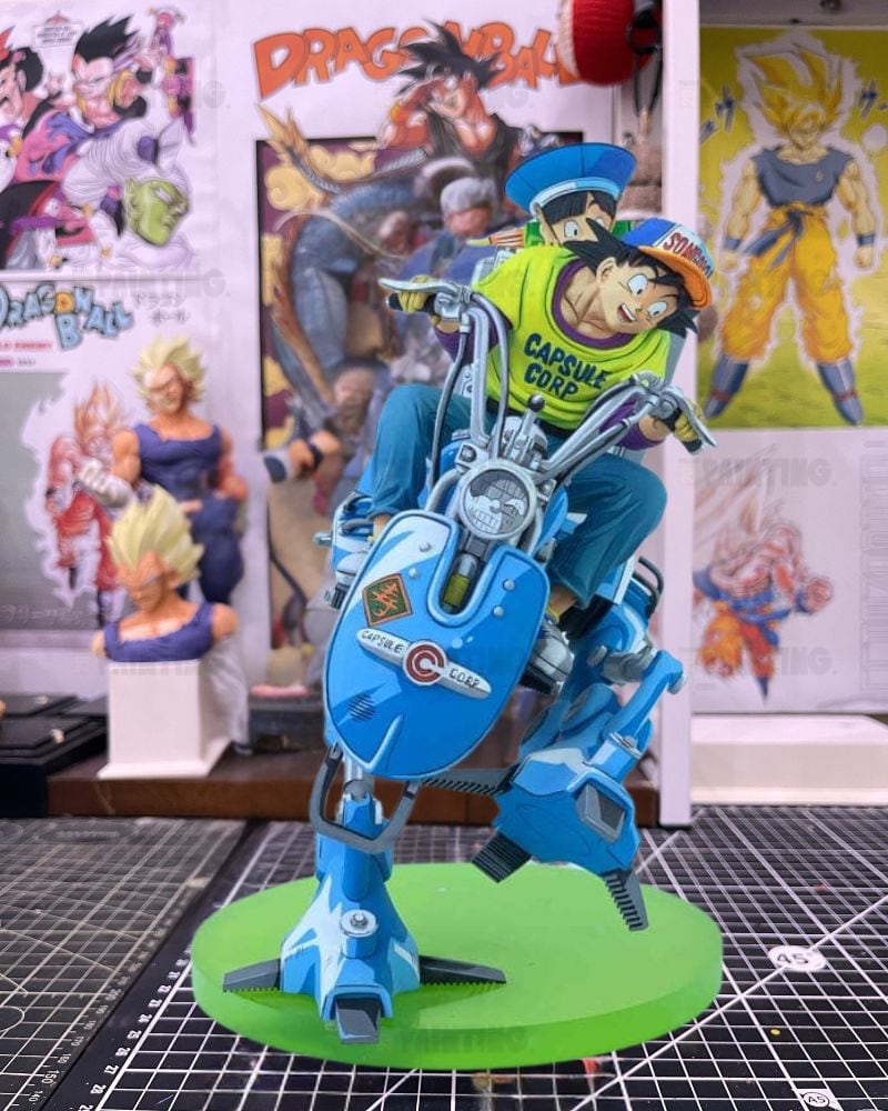 A detailed collectible statue of a character riding a blue motorcycle-like vehicle on a green circular base, set against a background of Dragon Ball posters and other figurines.