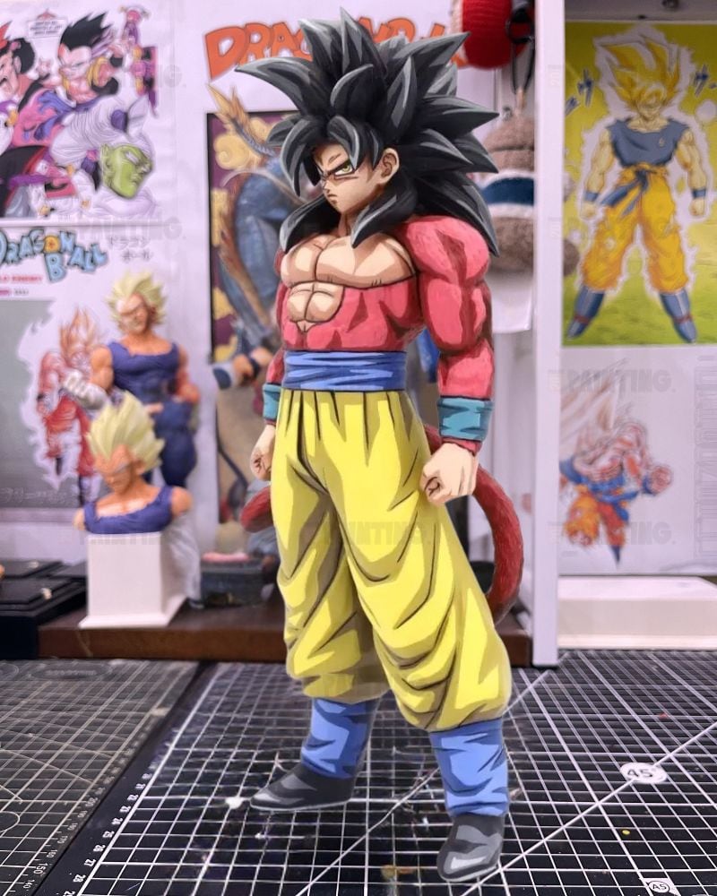 A detailed collectible figure of Goku in his Super Saiyan form, standing on a gridded cutting mat in a collector's room with Dragon Ball posters and figurines in the background.