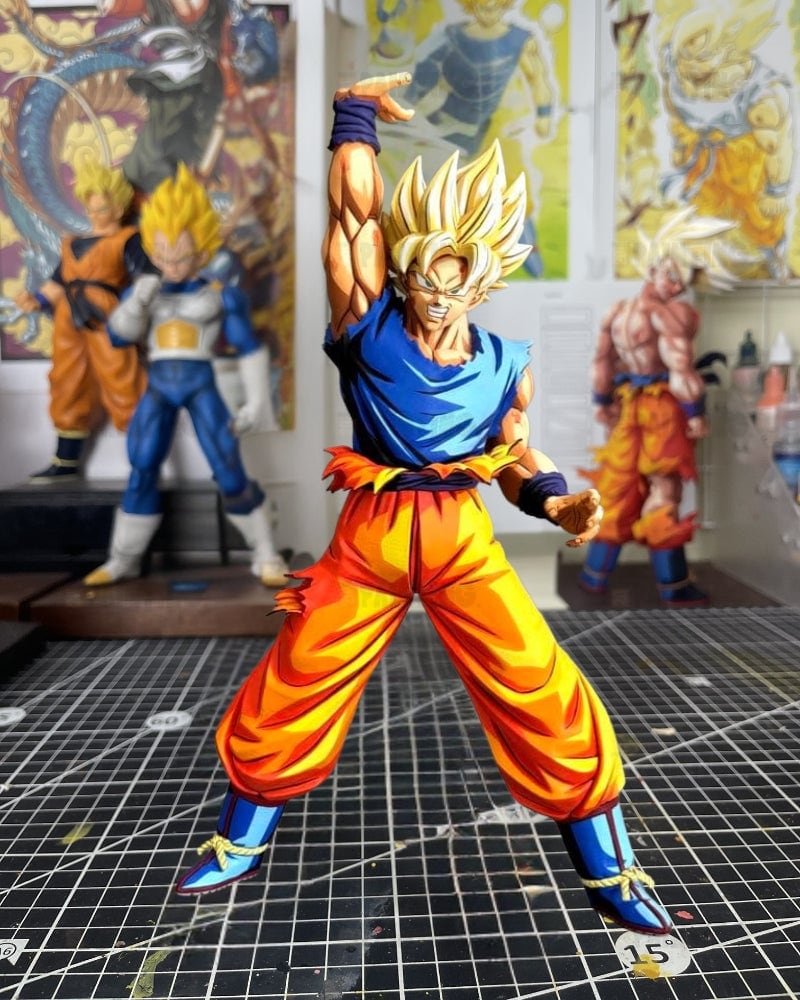A detailed collectible action figure of Goku in a Super Saiyan pose, standing on a black gridded cutting mat in a workshop or hobby room with other anime figurines and posters in the background.