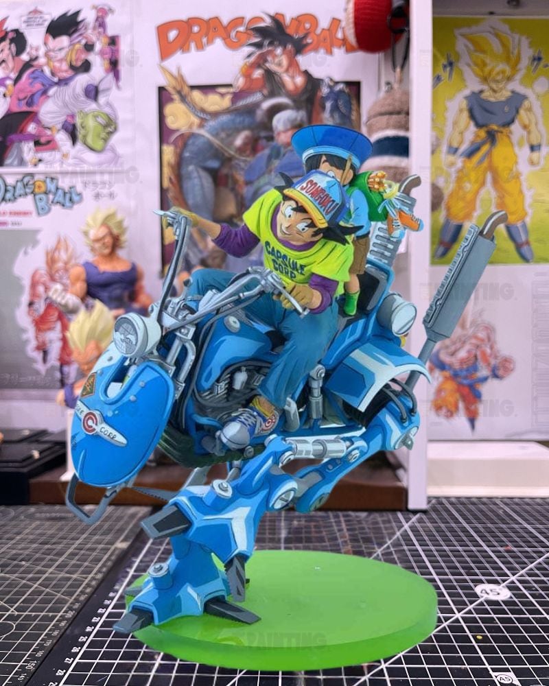 A detailed collectible figurine of a character riding a blue robotic motorcycle, mounted on a green circular base. The background shows anime posters on a shelf in a room setting.