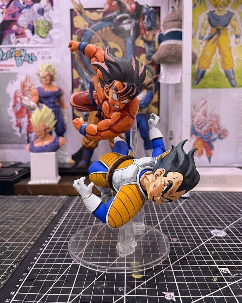 Dragon Ball figure set featuring Goku and Vegeta in dynamic combat poses on a clear acrylic stand, displayed on a cutting mat with Dragon Ball posters in the background.