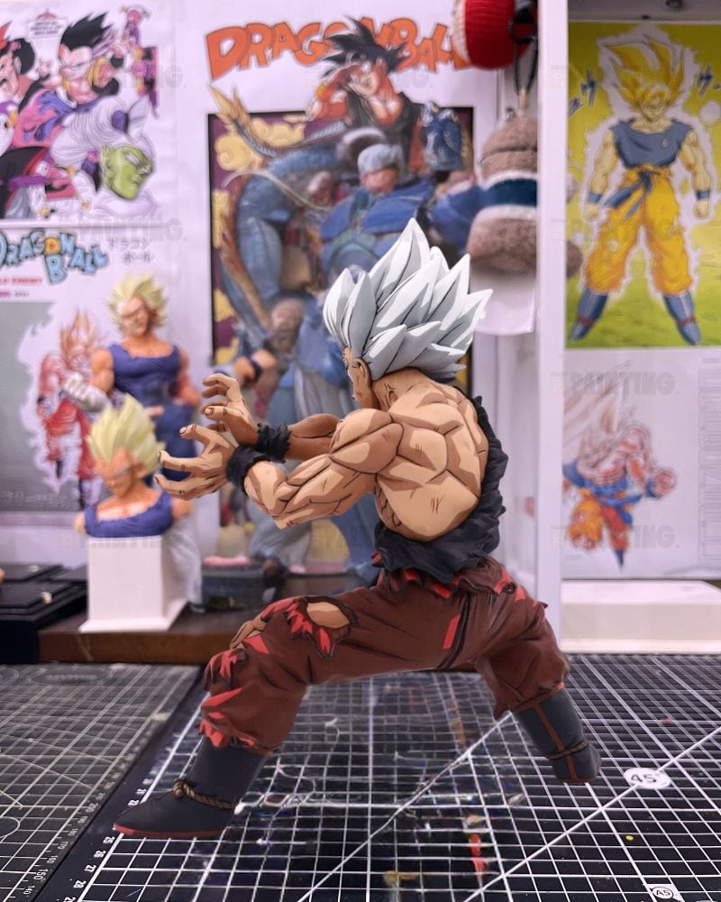 A detailed collectible action figure of Goku in a battle stance, featuring spiky silver hair, a torn black top, and torn brown pants, displayed on a gridded cutting mat with Dragon Ball posters in the background.