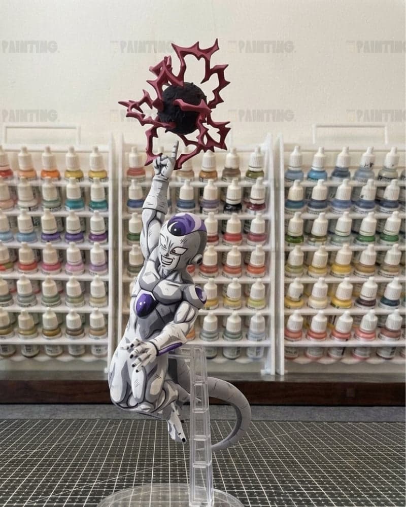 A painted anime figurine of Frieza from Dragon Ball Z, holding a red spiky orb, mounted on a clear stand in front of shelves of paint bottles.