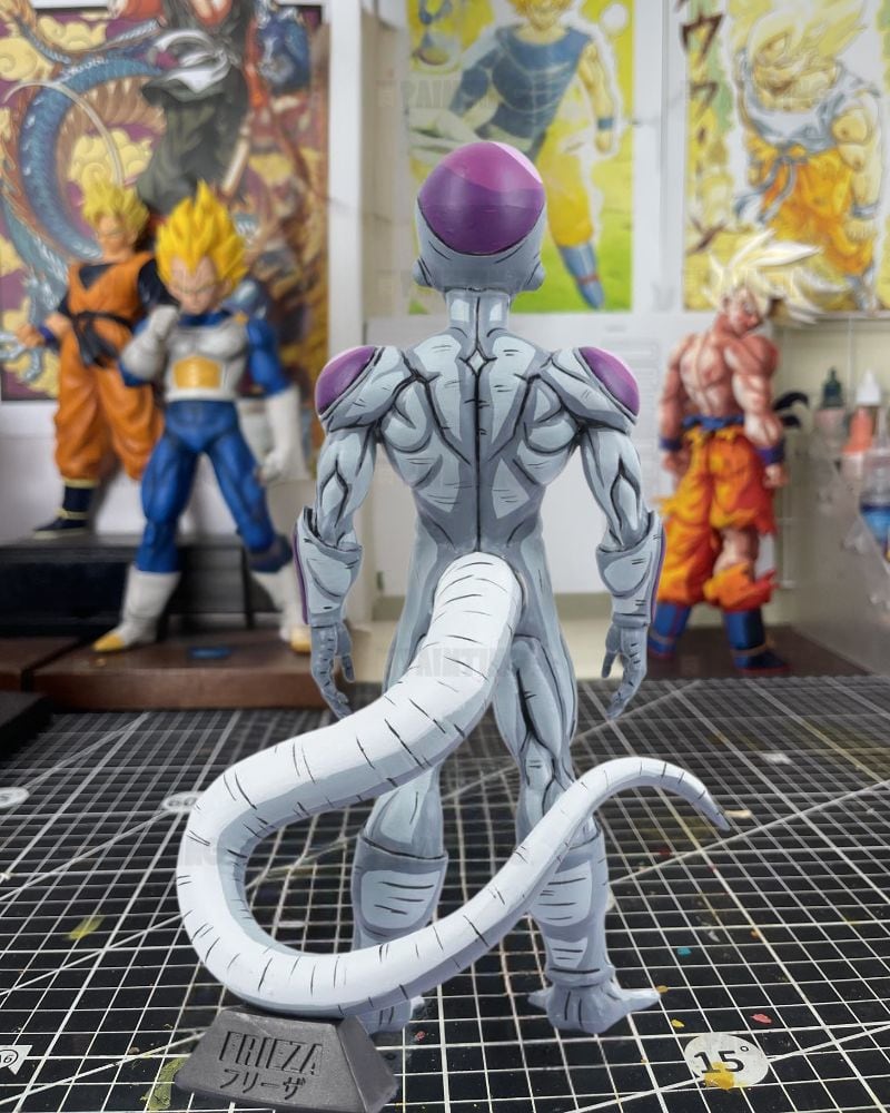 A detailed collectible figure of Frieza from Dragon Ball Z, shown from the back on a grid cutting mat with other anime figures in the background.