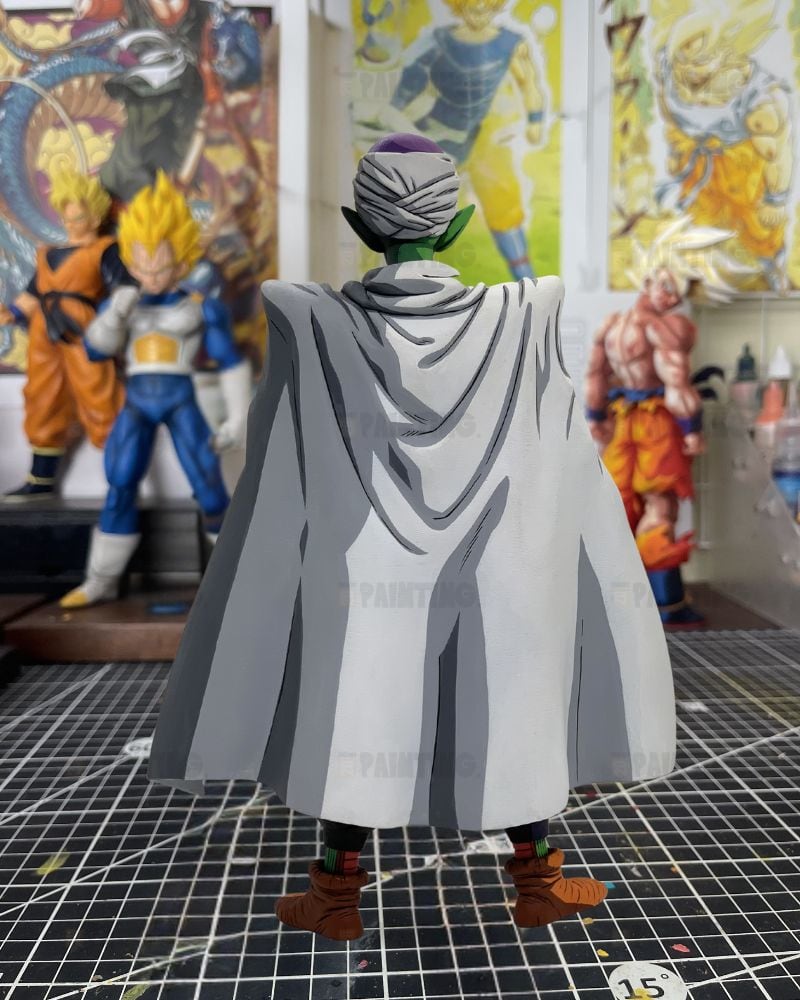 A Dragon Ball figure of Frieza from behind, wearing his signature gray cloak and green skin, standing on a gridded cutting mat with collectible figures in the background.