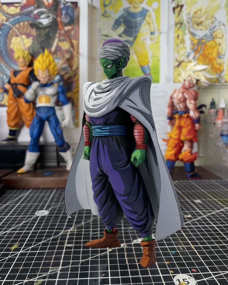 A Dragon Ball Z collectible figure of Piccolo standing on a gridded cutting mat, with other anime figures and posters blurred in the background.