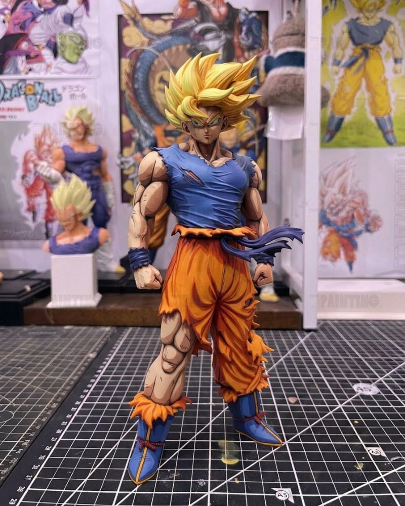 A detailed anime figurine of Goku in a Super Saiyan transformation, standing on a black gridded cutting mat with Dragon Ball collectibles in the background.
