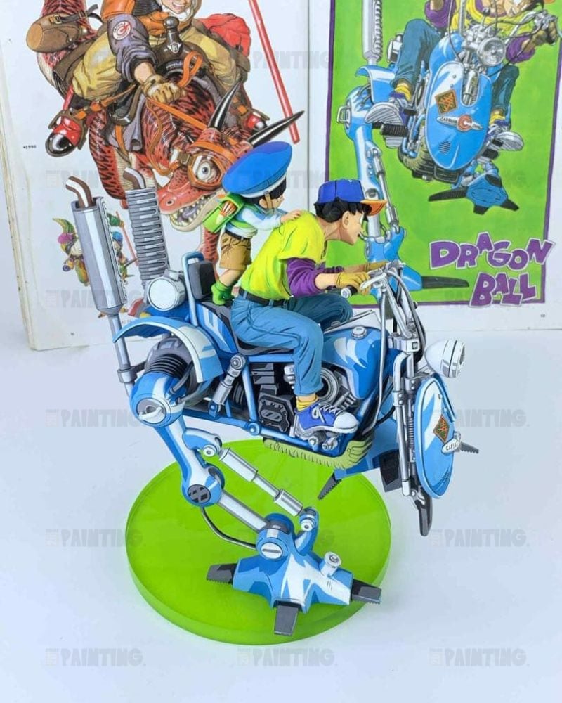 A detailed collectible model of two children riding a blue and silver robotic motorcycle on a green circular base, displayed in front of Dragon Ball themed posters.