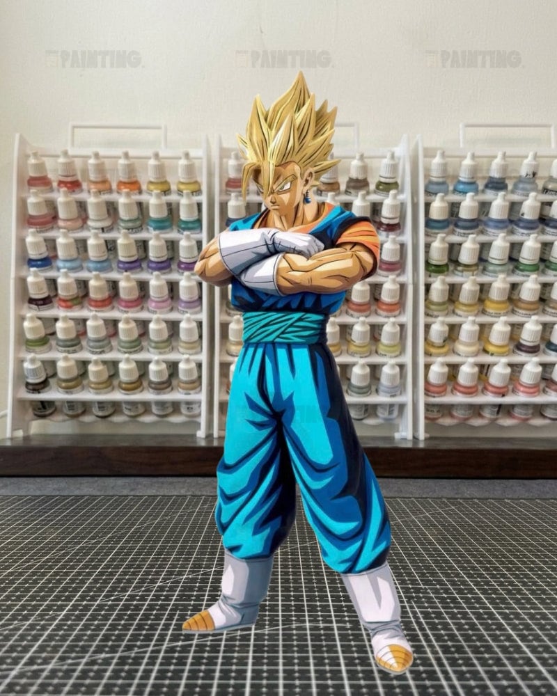 A die-cut sticker or decal of the anime character Vegetta from Dragon Ball Z, standing on a black gridded cutting mat with shelves of paint bottles in the background.