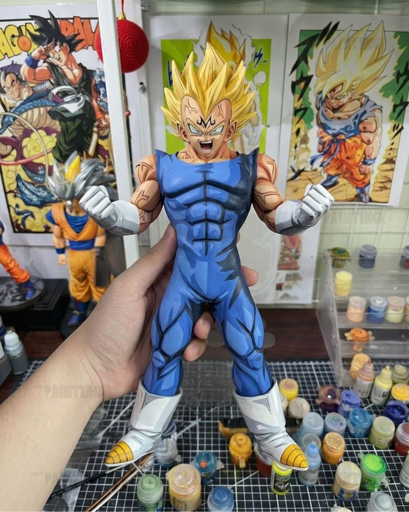 A hand holds a painted Dragon Ball Z Super Saiyan Gohan action figure in a blue jumpsuit with white gloves and boots, set against a background of anime posters and paint bottles.