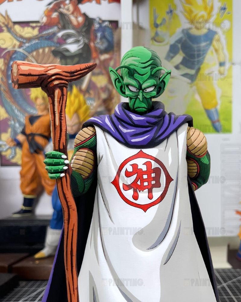 A detailed collectible figure of the Dragon Ball Z character Piccolo Sempai, featuring his signature green skin, purple collar, white robe with a red Chi symbol, and holding a wooden staff. The figure is posed in front of a wall filled with colorful Dragon Ball anime posters.
