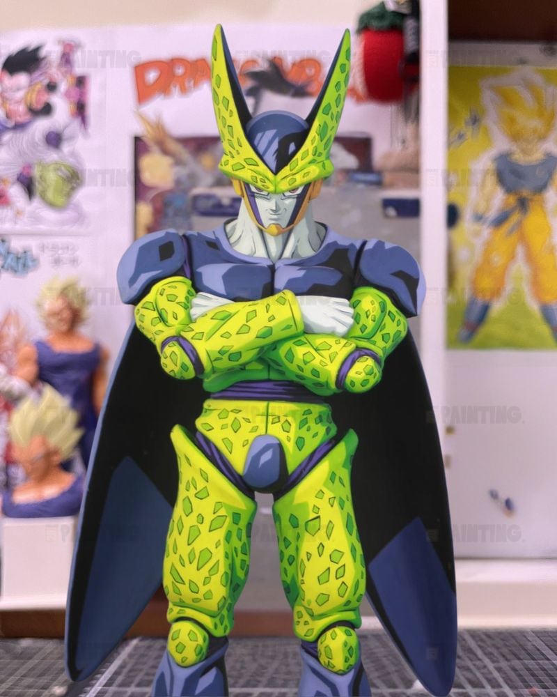 A Dragon Ball figure of Cell with arms crossed, featuring green and yellow patterned armor, a dark blue cape, and a blue helmet, set against a background of anime posters.