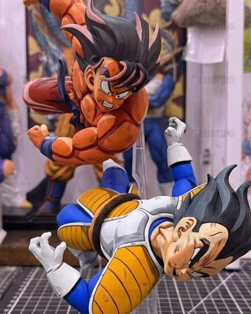 A detailed collectible figurine set featuring two anime characters in dynamic combat poses, one in an orange gi and one in a white and blue gi, displayed on a grid floor with other figures blurred in the background.