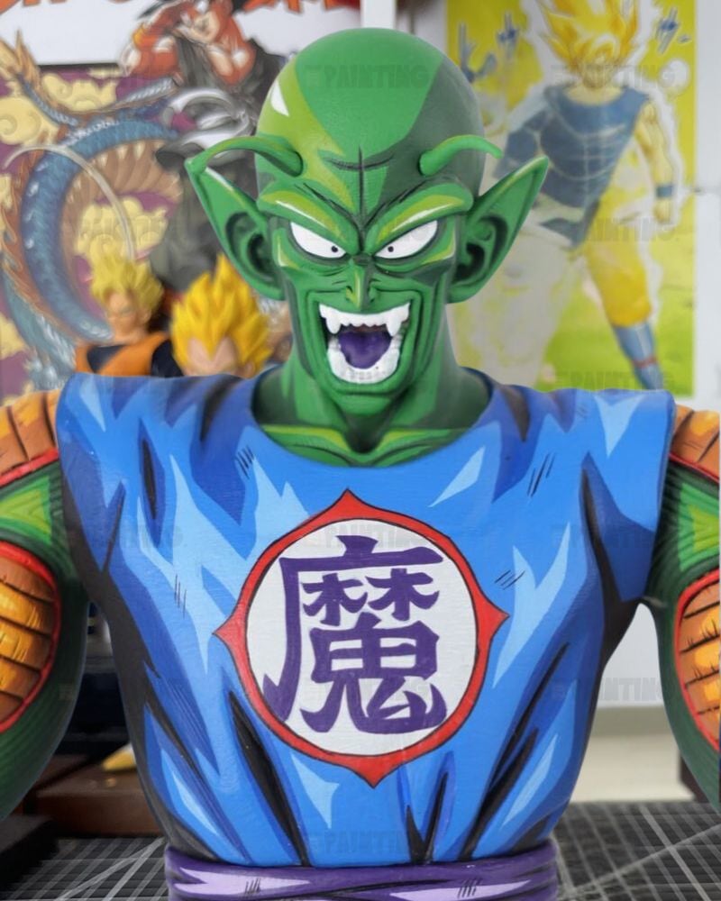 A collectible bust statue of Piccolo from Dragon Ball Z, featuring his signature green skin and blue gi with the Dragon Ball symbol on the chest, set against a blurred background of Dragon Ball posters.