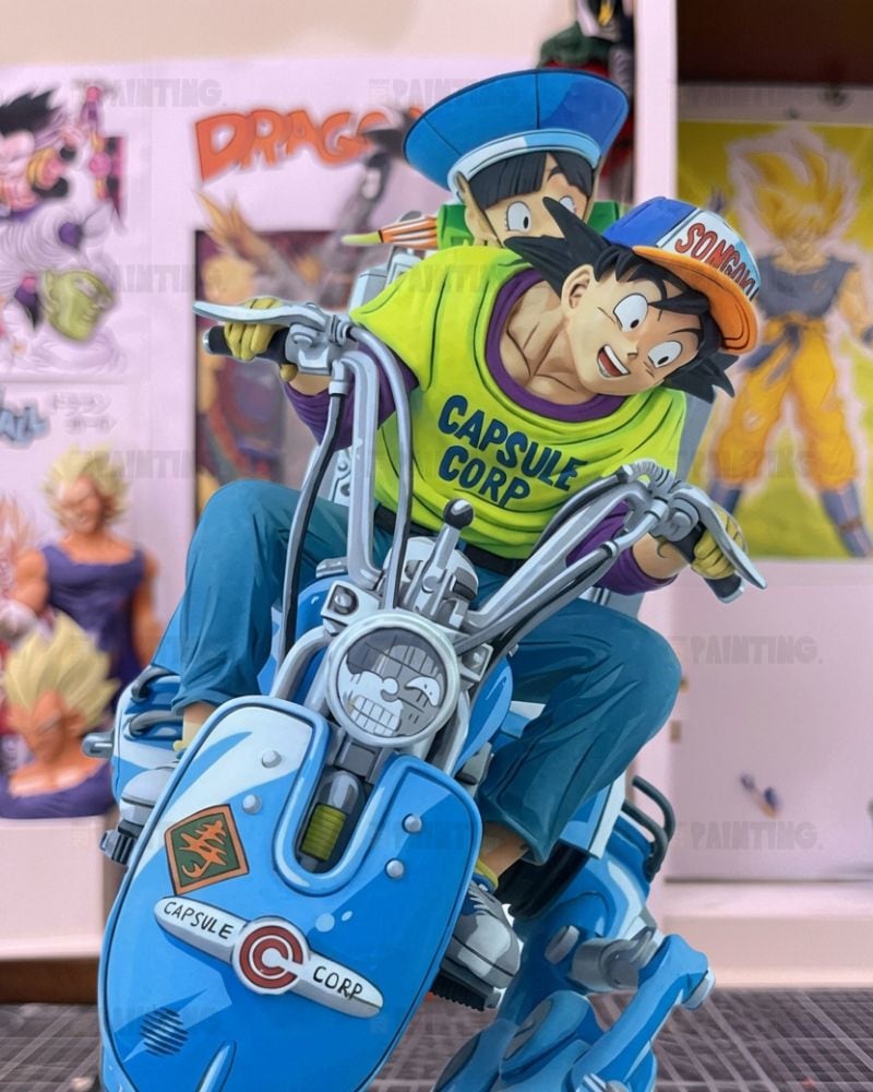 A collectible anime figurine of Goku and Vegeta riding a blue Capsule Corp motorcycle, displayed in front of a wall with Dragon Ball posters.