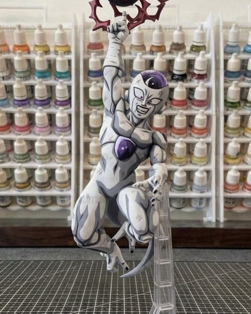 A painted anime figurine of a white and gray female character with purple accents, holding a red weapon, posed on a clear stand in front of shelves of miniature paint bottles.