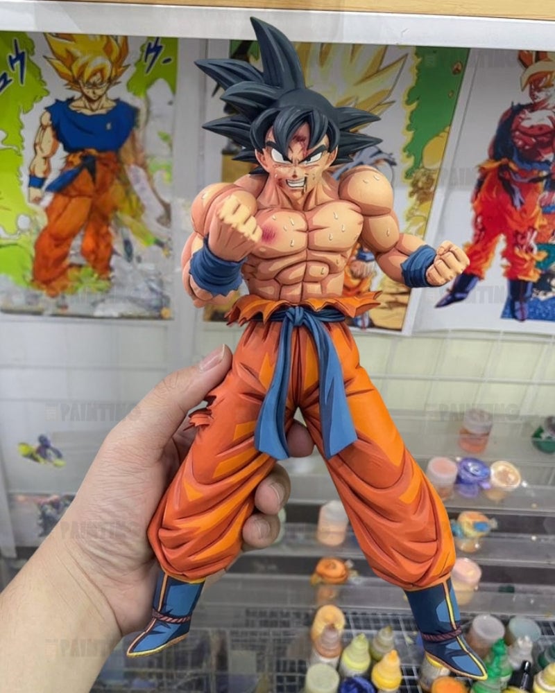 A hand holds a detailed anime figurine of Goku from Dragon Ball Z, wearing his signature orange gi and blue sash, with posters of Goku in the background.
