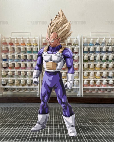 An anime-style collectible figure of Vegeta from Dragon Ball Z, standing on a cutting mat with shelves of paint bottles in the background.
