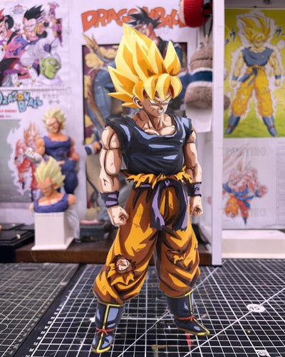 A detailed Dragon Ball figure of Goku in Super Saiyan form, standing on a gridded cutting mat with Dragon Ball posters and other collectibles in the background.