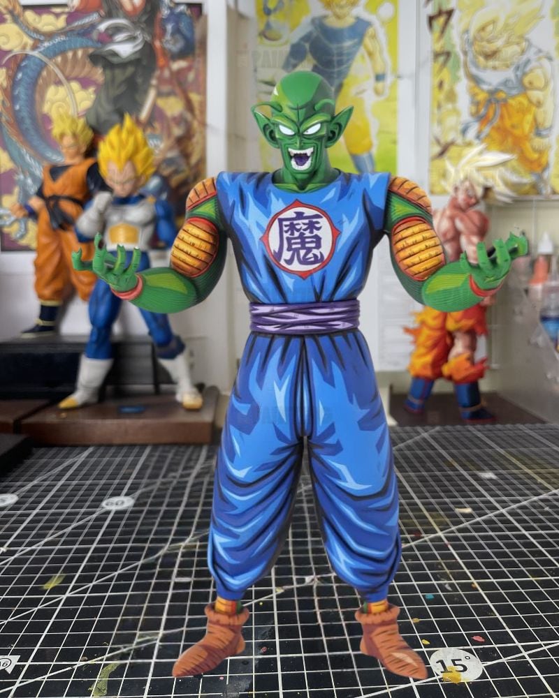 A detailed collectible action figure of Piccolo from Dragon Ball Z standing on a black gridded cutting mat, with other Dragon Ball figures blurred in the background of a room.