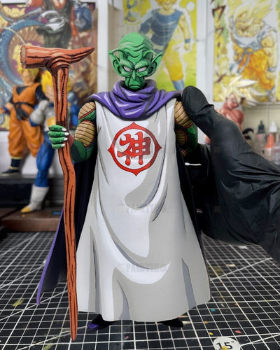A detailed action figure of Piccolo from Dragon Ball Z, featuring his green skin, white robe with a red Chi symbol, purple cape, and wooden staff, being posed by a hand wearing a black glove in a collector's display room.
