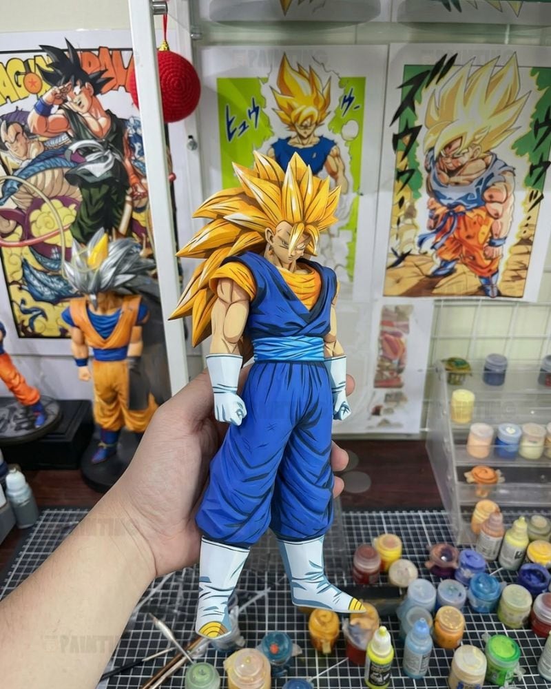 A hand holds a detailed anime figurine of Goku in a Super Saiyan form from Dragon Ball, wearing his blue and orange gi with white gloves and boots. The background shows a cluttered hobby room with Dragon Ball posters, a smaller Goku figure, and paint bottles on a table.