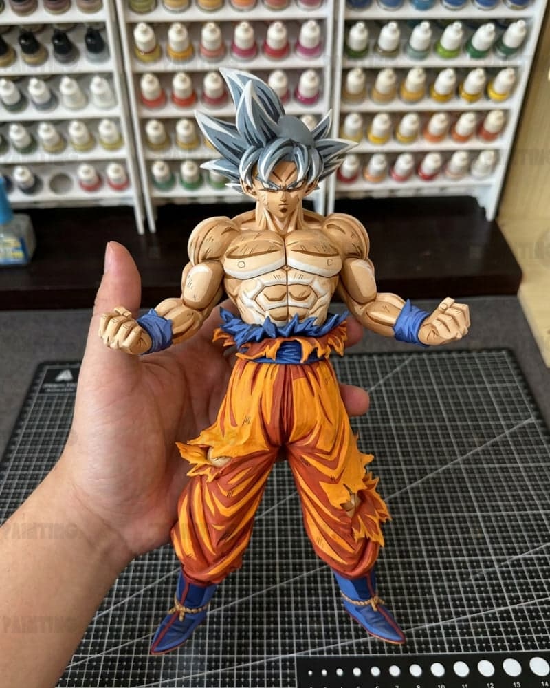 A hand holds a detailed anime figurine of Goku in his Super Saiyan form, featuring spiky silver hair, a muscular tan body, torn orange pants, and blue wristbands and boots, set against a background of shelves filled with small bottles.