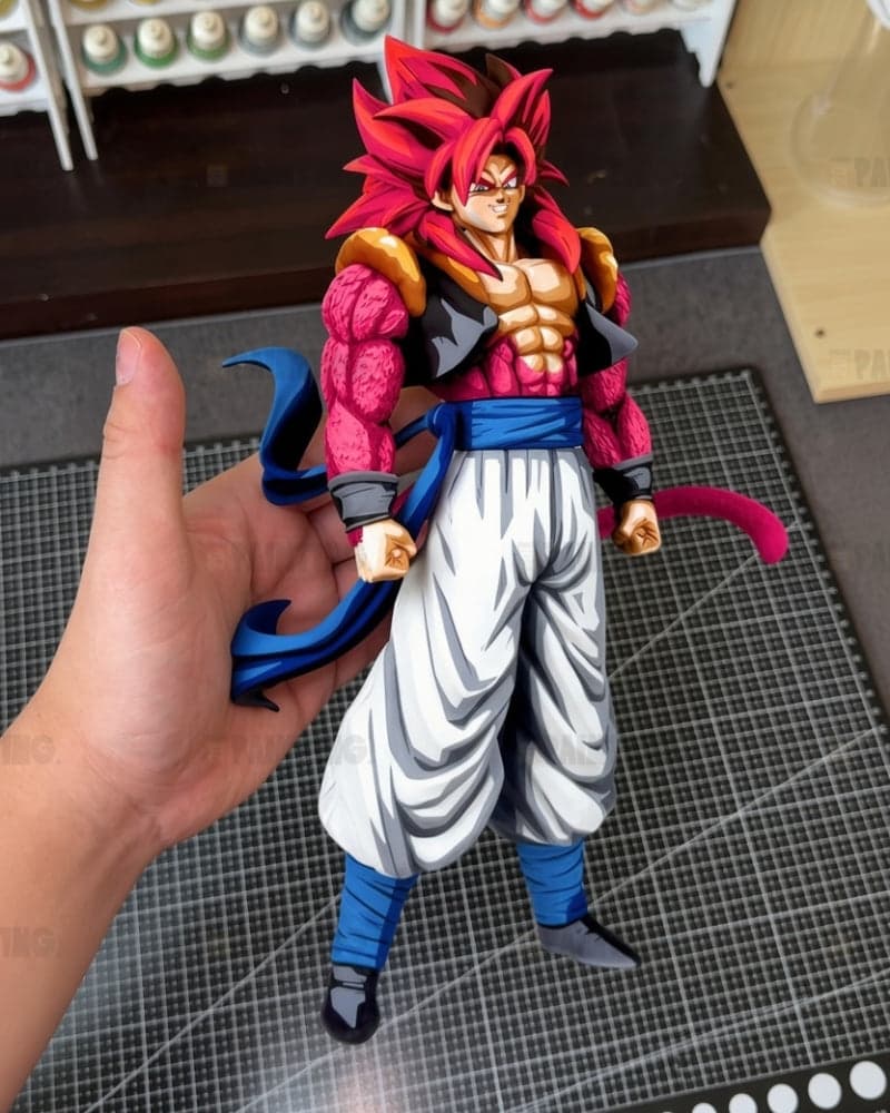 A hand holds a detailed anime figurine resembling Super Saiyan Gohan from Dragon Ball, featuring spiky red hair, a pink and black bodysuit, white pants, and blue accents, set against a tabletop background with a gridded mat.