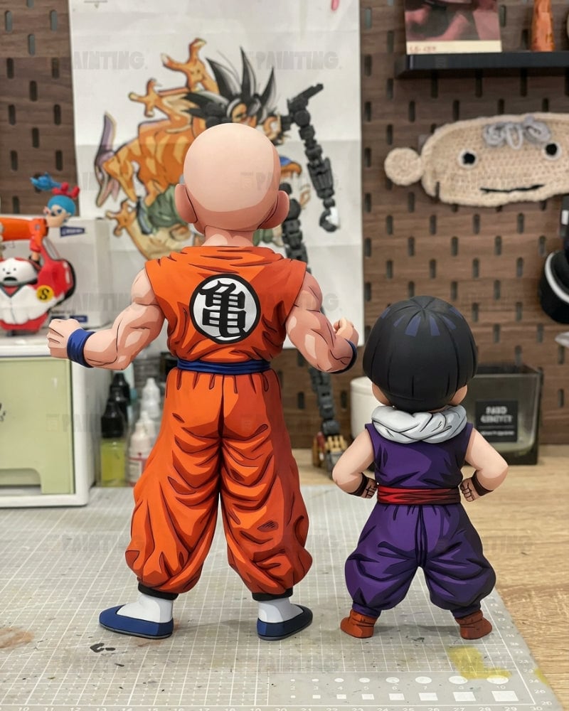 Two Dragon Ball Z action figures shown from the back, one resembling Goku in an orange gi and the other resembling Vegeta in a purple gi, standing on a cutting mat in a workshop setting.