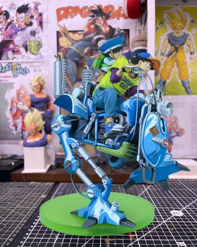A detailed Dragon Ball Z anime figurine of Goku and Vegeta riding a blue mechanical motorcycle on a green circular base, set against a background of Dragon Ball posters and other collectibles.