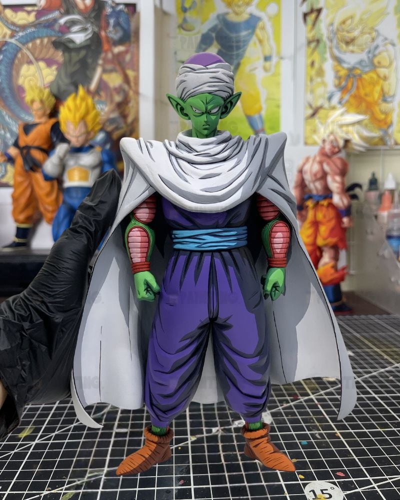 A detailed anime action figure of Piccolo from Dragon Ball Z, standing on a gridded cutting mat with a black-handled tool on the left and other collectible figures in the background.