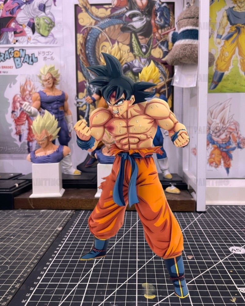A detailed anime figurine of Goku in a fighting pose, standing on a gridded cutting mat with Dragon Ball posters and other collectibles in the background.