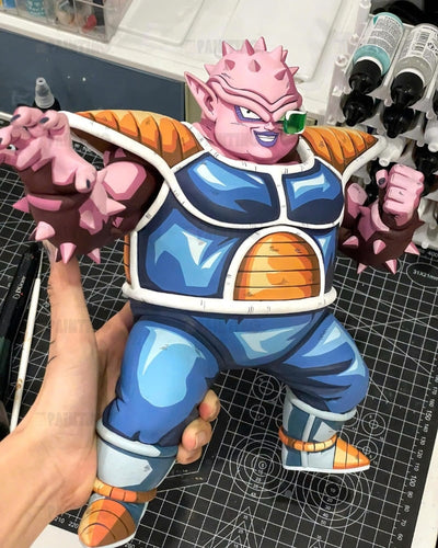 A hand-painted collectible figurine of a pink-skinned Dragon Ball Z character with spiky hair, wearing a blue and orange armor suit, held up by a person's hand over a cutting mat.