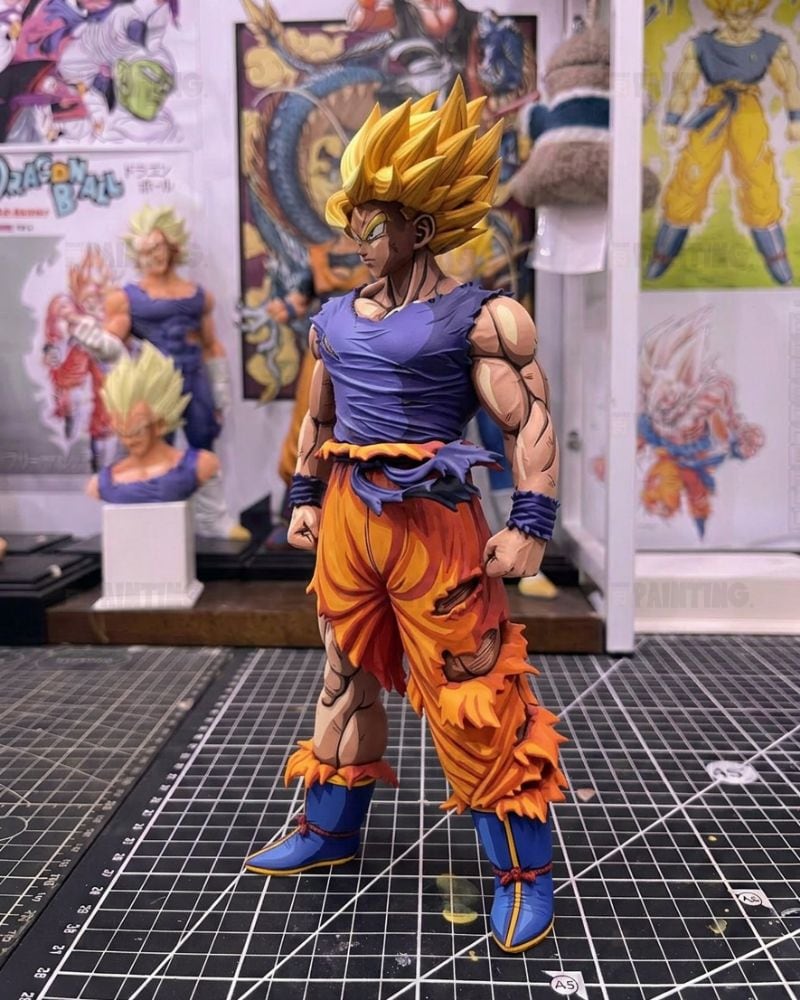 A detailed collectible action figure of Goku in a Super Saiyan form, standing on a black gridded cutting mat with Dragon Ball Z themed posters and other figurines in the background.