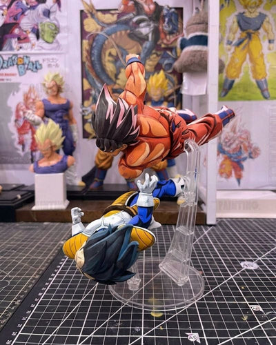 A detailed collectible action figure display featuring Goku in an orange gi performing a flying kick on Vegeta, who is lying on a clear acrylic stand. The figures are set on a black gridded cutting mat with Dragon Ball Z posters in the background.