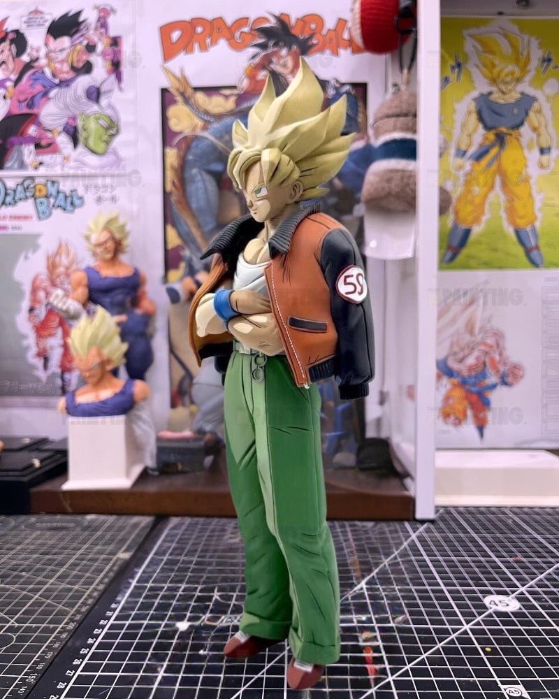 A Dragon Ball figure of a Super Saiyan with spiky blond hair, wearing a brown and black jacket, white undershirt, and green pants, standing on a black gridded cutting mat in a collector's room with Dragon Ball posters in the background.
