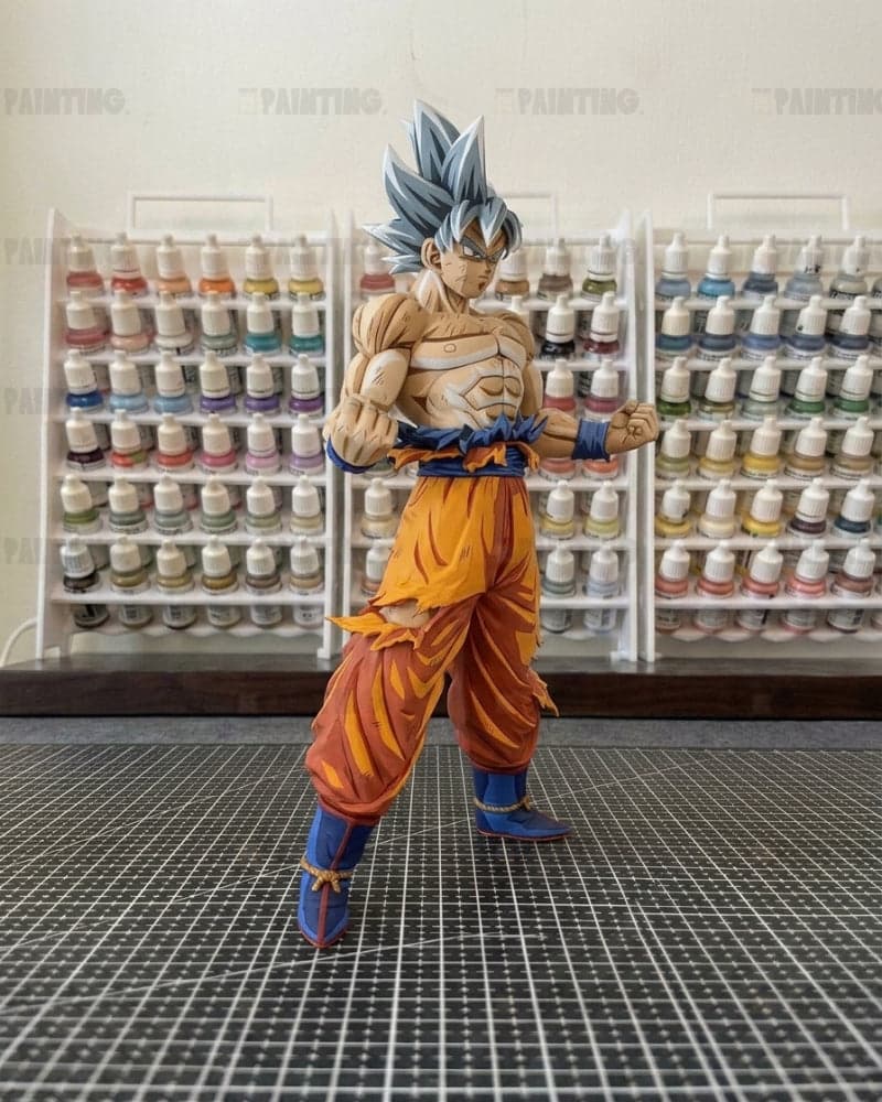 A detailed anime figurine of Goku in a blue-haired Super Saiyan form, standing on a black grid mat with shelves of paint bottles in the background.