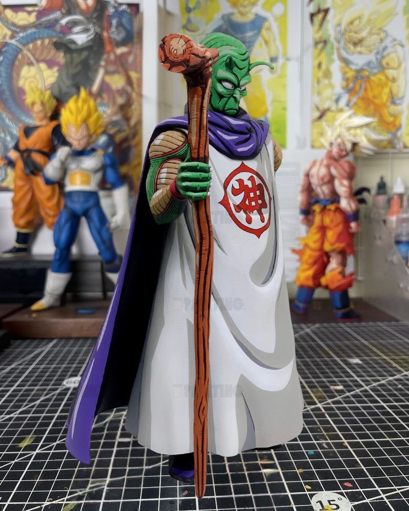 A detailed collectible figurine of a green-skinned character from Dragon Ball Z, wearing a white robe with a red symbol on the chest and a purple cape, holding a wooden staff with a skull topper. The figurine is displayed on a gridded cutting mat with other anime figures blurred in the background.