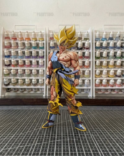 A detailed anime action figure of Goku in a Super Saiyan outfit, standing on a black gridded cutting mat with shelves of paint bottles in the background.