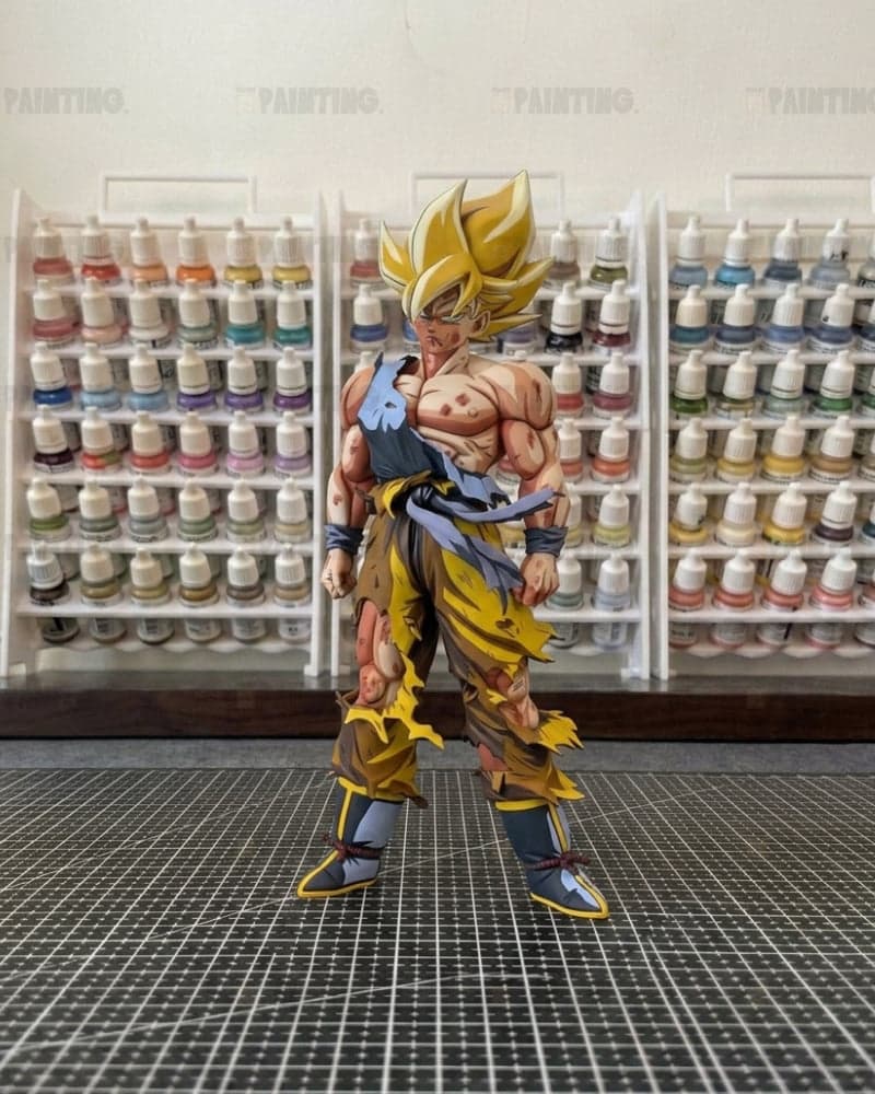A detailed anime action figure of Goku in a Super Saiyan outfit, standing on a black gridded cutting mat with shelves of paint bottles in the background.