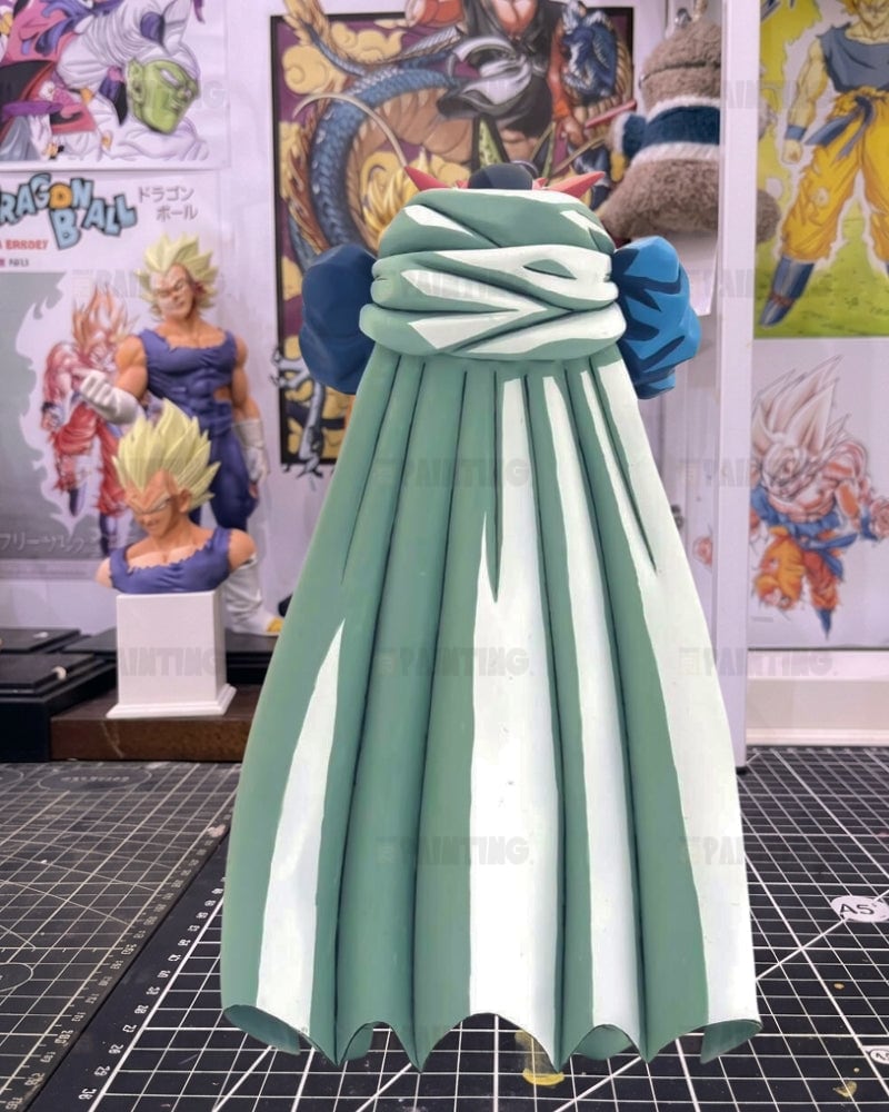 A miniature collectible figure of a character wearing a long pleated dress, shown from the back. The dress is sage green with white vertical pleats and a matching green and white criss-cross sash at the waist, flanked by blue shoulder accents.