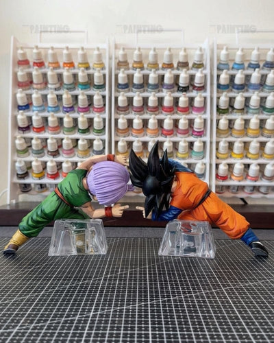 A collectible anime figurine of two characters in a dynamic fighting pose, displayed on a black gridded cutting mat with shelves of miniature paint bottles in the background.