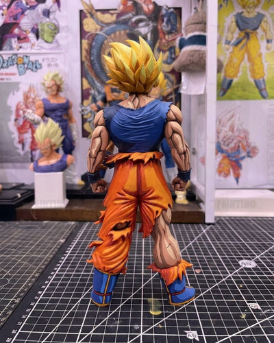 A detailed anime action figure of Goku in a Super Saiyan form, shown from the back on a black gridded cutting mat with other collectibles in the background.