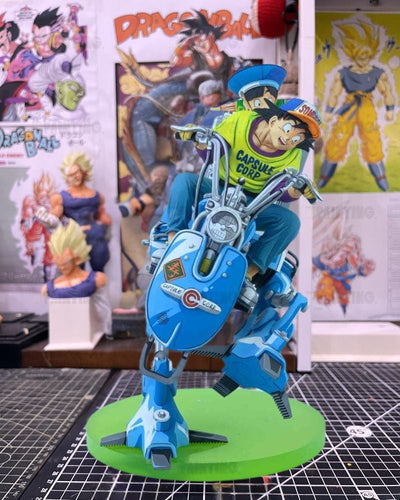 A detailed collectible statue of a character riding a blue motorcycle-like vehicle on a green circular base, set against a background of Dragon Ball posters and other figurines.